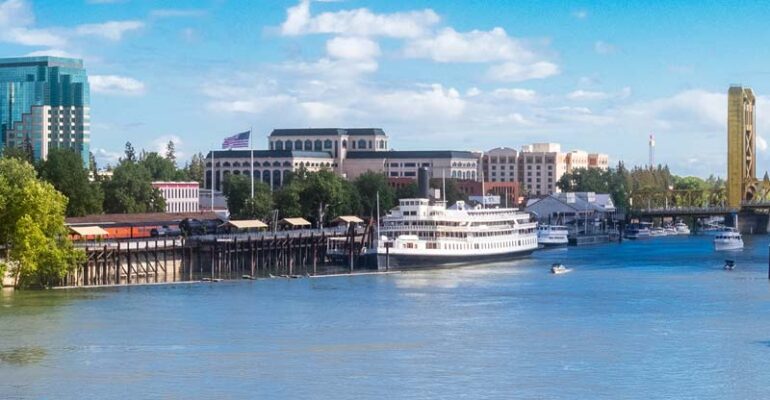 A panoramic view of Sacramento River near Old Town Sacramento, CA. A large ship is in the doc on the river near a bridge.