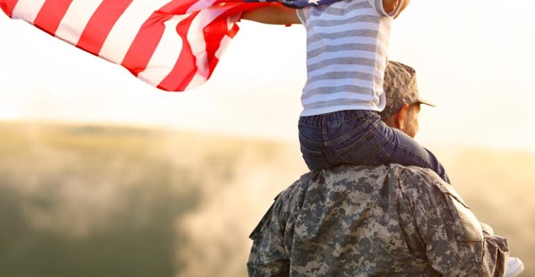 A Veteran with a young child on his shoulders holding an American Flag blowing in the wind.