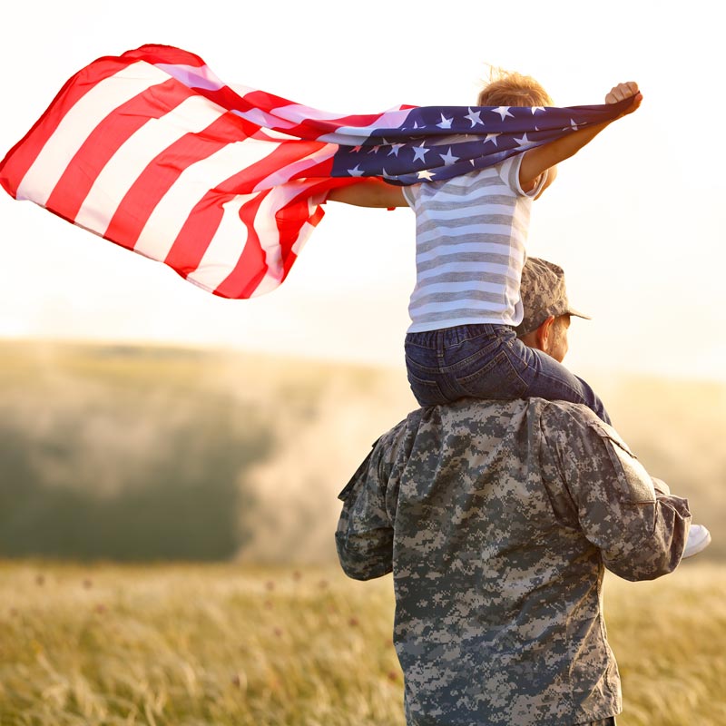 A Veteran with a young child on his shoulders holding an American Flag blowing in the wind.