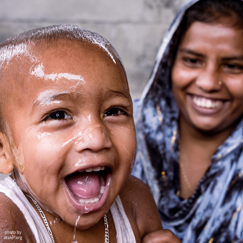 This image shows a mother and her baby smiling and laughing together, while the baby is soaked with water.