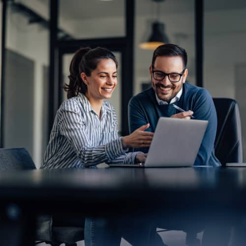 Two professionals smile while talking about what they're seeing on a laptop in an office.